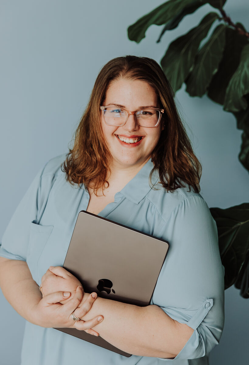Jenn McGeehan smiling, standing beside a fiddle leaf fig with her laptop